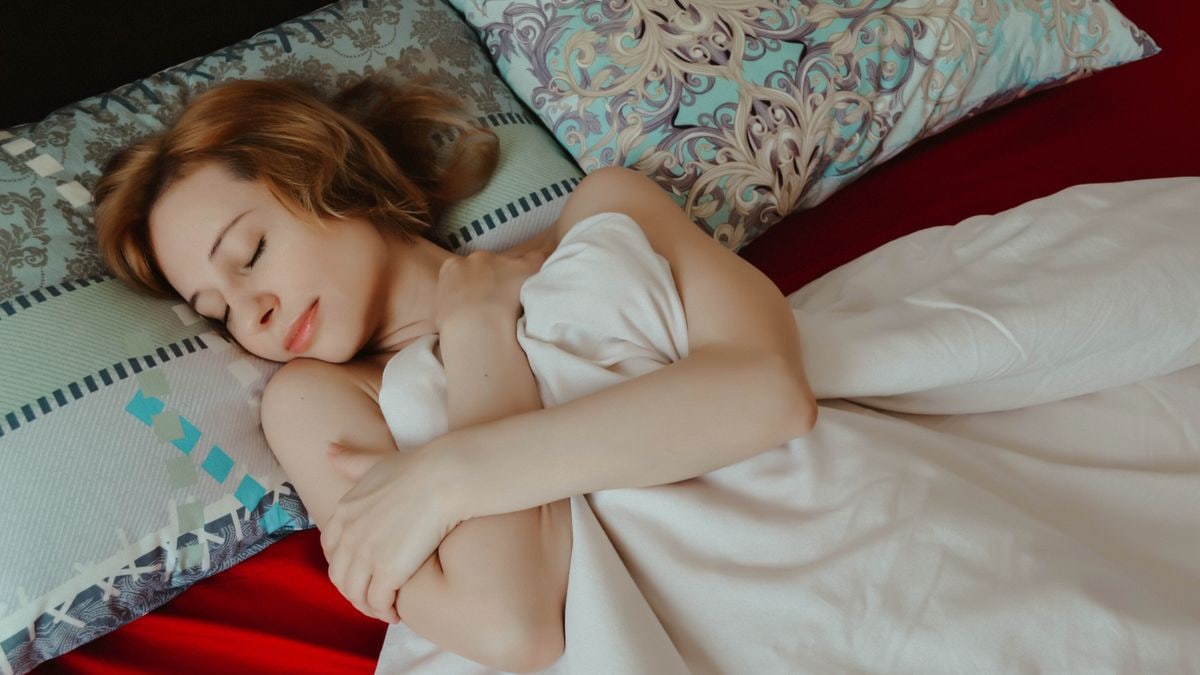 From above of relaxed female with arms crossed taking nap while lying on comfortable bed with pillows under blanket in bedroom